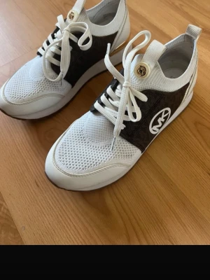 Vita och bruna Michael Kors sneakers - Snygga sneakers från Michael Kors i vitt och brunt med tydligt MK-logo på sidan. Ovandelen är i mesh och läderimitation, med vita snören och guldfärgade detaljer. Perfekta för dig som vill ha en sportig men ändå trendig look.
