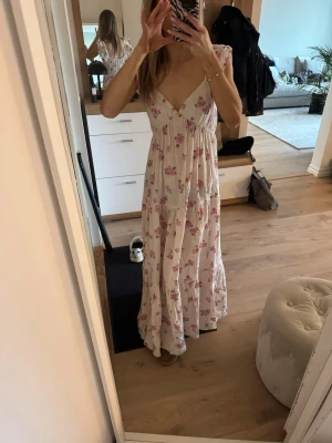 Vit långklänning med rosa blommor - Romantisk långklänning i vit bomull med rosa blommigt mönster. Klänningen har v-ringning, smala axelband och volangdetaljer vid axlarna. Perfekt för sommarens alla tillfällen och ger en luftig och feminin känsla.