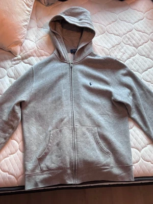 Grå zip-hoodie från Polo Ralph Lauren - Snygg grå hoodie med dragkedja från Polo Ralph Lauren. Klassisk design med huva, två fickor framtill och liten broderad logga på bröstet.                Storlek: XL (18-20)
