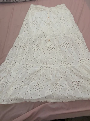 Vit långkjol med broderade detaljer - Superfin vit långkjol med broderade hålmönster och volanger. Kjolen har en elastisk midja med snörning och små träpärlor som detalj. Perfekt för varma dagar och ger en romantisk boho-vibe till din outfit.