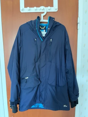 Warp TCS 3000 jacket - Mörkblå regnjacka från Warp med huva, flera fickor med dragkedja och justerbara ärmslut. Jackan har blått innerfoder och detaljer i turkos. Materialet är vattenavvisande TCS Water 3000, perfekt för regniga dagar.
