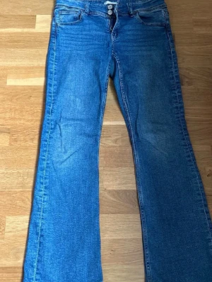 Blå bootcut jeans med fickdetaljer - Ett par klassiska blå bootcut jeans med två knappar i midjan och fickor både fram och bak. Jeansen har en lätt utsvängd passform och är tillverkade i denim med snygga sömdetaljer. Perfekta för en avslappnad och trendig look. Lite slitna längst nere vid benen 