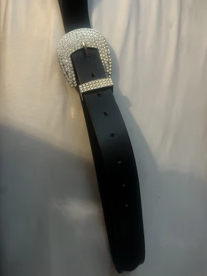 Svart bälte med glittrigt spänne - Stilrent svart bälte med ett stort, glittrigt silverfärgat spänne täckt av gnistrande stenar. Perfekt för att lyfta en outfit och ge den extra edge. Bältet har flera hål för justering och är tillverkat i syntetmaterial.
