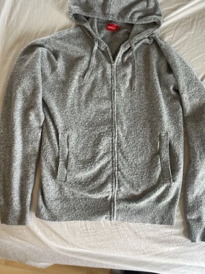 Grå hoodie med dragkedja  - Mysig grå hoodie med huva och snörning. Hoodien har dragkedja framtill och två fickor på sidorna. Perfekt för en chill och avslappnad stil. Tillverkad i mjukt material som känns skönt mot huden.