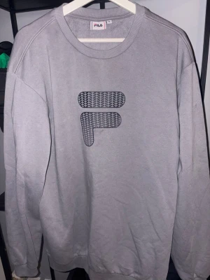 Grå sweatshirt från FILA - Grå sweatshirt från FILA med stort F-logga framtill i svart mönster. Tröjan har rund halsringning, långa ärmar och ribbade muddar vid ärmslut och nederkant. Perfekt för en avslappnad och sportig stil. Helt ny