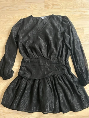Svart omlottklänning med struktur Klänning GinaStl 170 50kr - Snygg svart kortklänning med omlottliv och markerad midja. Klänningen har långärmade puffärmar och ett subtilt glittrigt mönster i tyget. Kjolen är lätt utställd och materialet känns mjukt och följsamt. Perfekt för dig som gillar trendiga detaljer.