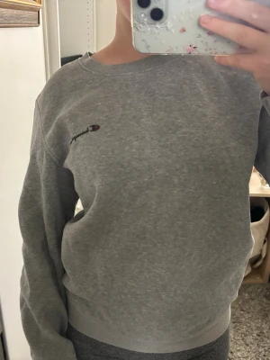 Grå sweatshirt från Champion - En klassisk grå sweatshirt från Champion med rund halsringning och broderad logga på bröstet. Tröjan har en avslappnad passform och är tillverkad i mjukt bomullsmaterial. Perfekt för en chill och sportig look.