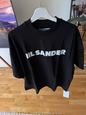 Svart Jil S. t-shirt med tryck - Fetaste T-shirt men den är oversize på mig säljer vidare. Large känns som xl men de är så med passformen .Cool svart t-shirt från Jil S. med vit logotyp framtill. Oversized passform och ribbad rund halsringning. Tillverkad i mjuk bomull som känns skön mot huden. Perfekt för en stilren och trendig look.