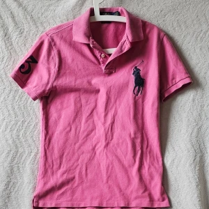 Rosa pikétröja från Ralph Lauren - Snygg rosa pikétröja från Ralph Lauren med klassisk krage, korta ärmar och stor marinblå logga på bröstet. Tröjan har även en broderad siffra på ena ärmen och är i en slim fit-modell. Perfekt för en sportig och stilren look.