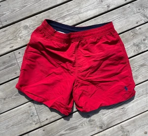 Röda badbyxor från Polo Ralph Lauren - Snygga röda badbyxor från Polo Ralph Lauren med klassisk marinblå logga broderad på ena benet. De har elastisk midja med dragsko, innerbyxa i mesh och en bakficka med lock. Perfekta för stranden eller poolen.