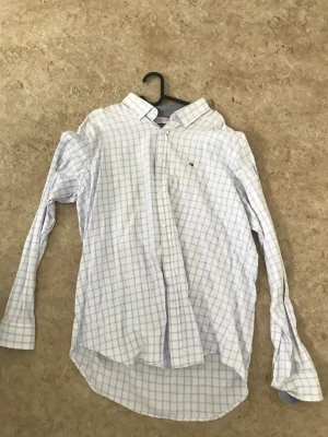 Vit rutig skjorta från Tommy Hilfiger - Vit skjorta med ljusblått rutmönster från Tommy Hilfiger. Klassisk krage, långa ärmar och knäppning framtill. Liten logga broderad på bröstet. Perfekt för en clean och stilren look.