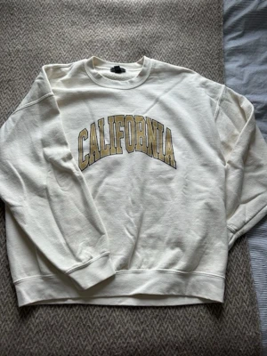 Beige sweatshirt med CALIFORNIA-tryck - Snygg beige sweatshirt med stort broderat CALIFORNIA-tryck i beige och svart på bröstet. Klassisk rund halsringning och ribbade muddar vid ärmslut och nederkant. Perfekt för en avslappnad och trendig look.