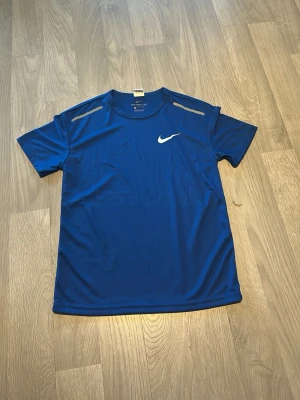Blå tränings-tshirt från Nike - Säljer en blå tränings-tshirt från Nike med vit logga på bröstet och reflekterande detaljer på axlarna. Tillverkad i lätt och ventilerande polyester, perfekt för träning och sportaktiviteter.