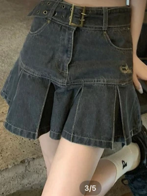 Svart jeanskjol med bälte och volang - Trendig svart jeanskjol med bredt bälte och guldspänne. Kjolen har volangdetaljer och markerade sömmar för en cool look. Perfekt för dig som gillar streetstyle och vill sticka ut.