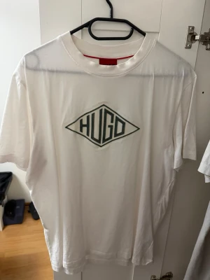 Vit t-shirt  Hugo  - Vit t-shirt från Hugo med stor svart logga på bröstet i grafisk stil. Klassisk rund halsringning och korta ärmar. Perfekt för en stilren och avslappnad look.