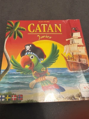 Catan Junior brädspel - Catan Junior är ett färgglatt brädspel med pirattema där spelare bygger på en övärld. Spelet har en papplåda med illustration av en papegoja med pirathatt, skepp och palmer. Perfekt för familjer och yngre spelare som gillar strategi och äventyr.