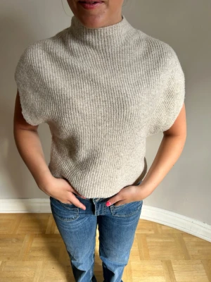 Beige ribbstickad ärmlös tröja💖 - Säljer en beige ribbstickad ärmlös tröja med hög hals. Tröjan har en avslappnad passform och är gjord i ett mjukt, tjockare material som känns lyxigt. Perfekt att styla över en skjorta eller t-shirt för en trendig lager-på-lager-look.