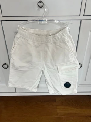 Vita shorts från C.P. Company 140  - Säljer ett par vita shorts från C.P. Company med elastisk midja och snörning. Shortsen har sidofickor och en stor ficka med lock och den klassiska C.P. Company-loggan på. Knappt använda