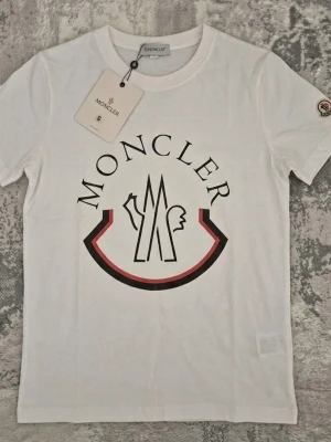 Vit Moncler t-shirt med logga - Vit t-shirt från Moncler med stor tryckt logga framtill i svart och rött. Klassisk rund halsringning och korta ärmar. Ikonisk Moncler-patch på ena ärmen. Perfekt för dig som gillar stilrena och exklusiva plagg.