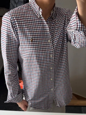 Rutig skjorta från Ralph Lauren - Klassisk rutig skjorta från Ralph Lauren i vitt, blått och rött. Skjortan har lång ärm, knappar framtill och broderad logga på bröstet. Perfekt för en stilren och avslappnad look. Den är lite missfärgad i kragen och armhålorna, därav priset. Annars bra skick 
