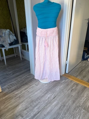 Ljusrosa lång kjol med smock - ljusrosa lång kjol med bred smock i midjan och dekorativ knytning framtill. perfekt nu till sommaren!💓tyvärr har några pärlor ramlat av på midje snörena, se sista bilden.