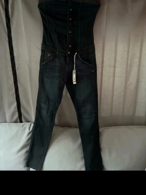 Mörkblå denim jumpsuit med hög midja - Cool mörkblå jumpsuit i denim med hög midja och knappstängning framtill. Snygga sömdetaljer och klassiska jeansfickor framtill. Perfekt för dig som vill sticka ut med en edgy look. Materialet är kraftigt denim och modellen är figurnära upptill.