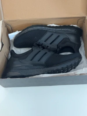 Svarta Adidas Ultraboost sneakers - Snygga helsvarta Adidas Ultraboost sneakers med stickad ovandel och klassiska tre ränder på sidan. Skorna har en chunky sula i Boost-material för extra komfort och en sportig look. Perfekta för dig som gillar stilrena och bekväma sneakers.