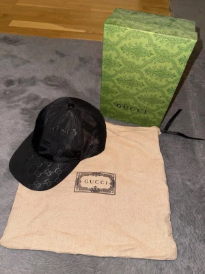 Svart keps från Gucci med GG-mönster - Snygg svart keps från Gucci med klassiskt GG-monogram över hela kepsen. Framsidan är i glansigt tyg och baksidan har mesh för extra ventilation. Kepsen levereras med originalpåse och låda. Perfekt accessoar för en trendig streetstyle.