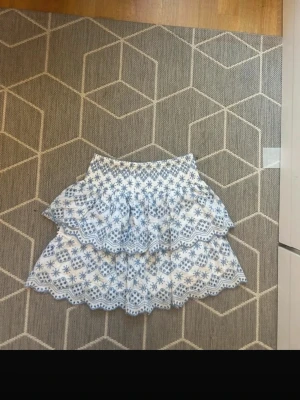 Vit och blå volangkjol med spets - Supersöt kort kjol i vitt och blått med volanger och spetsdetaljer. Kjolen har ett blommigt mönster och elastisk midja för skön passform. Perfekt för sommarens alla tillfällen!