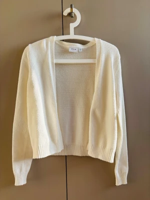 Vit öppen kofta från Vila - Stilren vit kofta från Vila med öppen front och långa ärmar. Koftan har ribbade muddar vid ärmslut och nederkant, vilket ger en snygg och enkel look. Perfekt att slänga över en topp för en avslappnad stil.