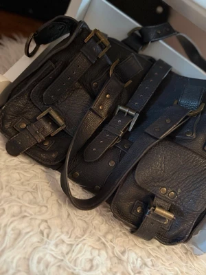 Mulberry Roxanne - Cool svart handväska i äkta läder med flera fickor och detaljerade mässingsspännen. Väskan har en robust och lite vintage look med justerbara remmar och många nitar. Perfekt för dig som gillar en edgy och praktisk stil.