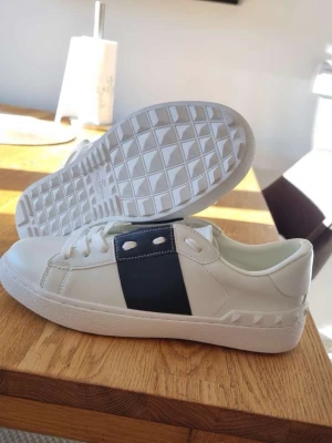 Valentino vita sneakers med svart detalj - Snygga vita sneakers från Valentino med en bred svart läderdetalj över snörningen och vita nitar på sidan. Klassisk låg modell med vit sula och diskret logga på insidan. Perfekta för dig som gillar stilrena och exklusiva skor.