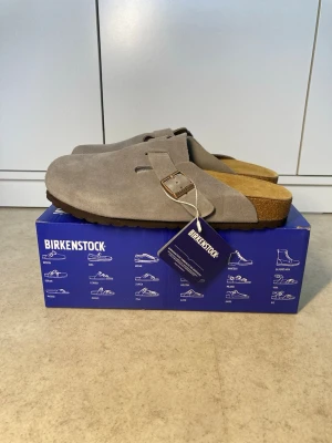 Birkenstock Boston tofflor grå mocka - Snygga Birkenstock Boston tofflor i mörkgrå mocka med klassisk design och justerbart spänne i metall. Fotbädden är i kork och sulan är mörkbrun. Perfekta för en avslappnad och trendig stil.