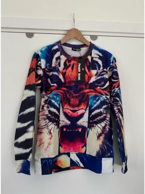 Färgstark college tröja med tigertryck - storlek S - Helt ny mönstrad collegetröja / sweatshirt från Peoples Focus i storlek S med ett stort, färgstarkt tigermotiv på framsidan. Ärmarna och baksidan har ett mönster som liknar tigerpäls i olika färger som blått, rött och svart. Klassisk modell med rund halsringning och muddar vid ärmslut och nederkant. Helt NY med etikett. 