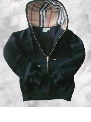 Svart hoodie från Burberry med dragkedja - Svart hoodie från Burberry med klassiskt rutigt foder i huvan och dragkedja framtill. Hoodien har ribbade muddar och snörning i huvan. Perfekt för dig som gillar stilrena och exklusiva plagg med en twist av ikoniskt mönster.