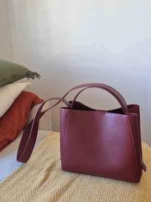 Vinröd handväska  - Snygg vinröd handväska i boxig modell med både handtag och lång axelrem. Väskan är gjord i läderimitation. Från Gina tricot, nästan helt oanvänd och mycket bra skick. Ca 25 cm x 11 cm x 22 cm. Minns ej nypris men runt 400 kr. 
