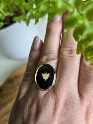 Svart ring med torkad blomma - Unik ring med en oval svart platta och en inbäddad torkad blomma i mitten. Ringen har guldfärgad metall och justerbart. Perfekt för dig som gillar naturinspirerade accessoarer och vill ha något personligt och stilrent.