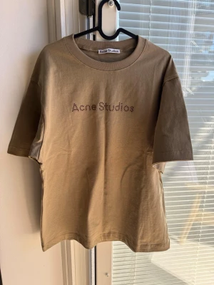Beige Acne Studios t-shirt med pärlor - Snygg beige t-shirt från Acne Studios med pärlbroderad logga på bröstet. Klassisk passform med rund hals och korta ärmar. Perfekt för dig som gillar stilrena plagg med en twist.