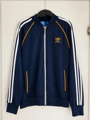Blå Adidas zip-tröja med ränder - Som ny ! Snygg blå zip-tröja från Adidas med klassiska vita ränder längs ärmarna och gula detaljer. Tröjan har dragkedja framtill, ribbade muddar och Adidas logga på bröstet. Perfekt för en sportig look.