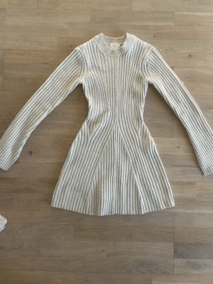 Beige ribbad stickad klänning - Superfin beige stickad klänning med ribbat mönster och lång ärm. Klänningen är kort och har en figurnära passform som framhäver midjan. Perfekt för dig som gillar en enkel men snygg stil.
