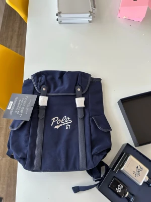 Mörkblå Polo 67 ryggsäck - Snygg mörkblå ryggsäck från Polo med broderad logga och siffran 67 på framsidan. Ryggsäcken har två sidofickor, justerbara axelband och detaljer i vitt. Tillverkad i slitstark canvas med praktisk design och klassisk look.