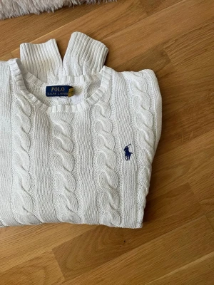 Vit kabelstickad tröja Polo Ralph Lauren - Vit kabelstickad tröja från Polo Ralph Lauren med rund halsringning och blå broderad logga på bröstet. Tröjan har långa ärmar och ribbstickade muddar. Perfekt för en klassisk och stilren look.