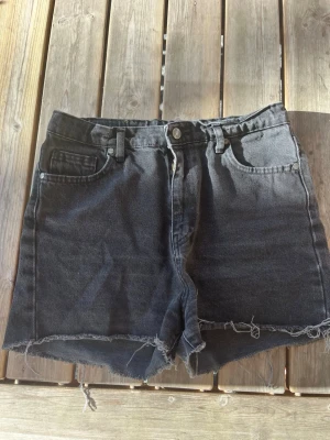 Svarta denimshorts med rå kant - Säljer ett par svarta högmidjade denimshorts från Denimday. Shortsen har fem fickor, gylf med dragkedja och råa, fransiga benslut för en avslappnad look. Midjan går att minska innanför shortsen men ena sidan är lite utsliten medan den andra knappt att man ser att den är använd. Perfekta till sommaren och lätta att matcha med olika stilar. 