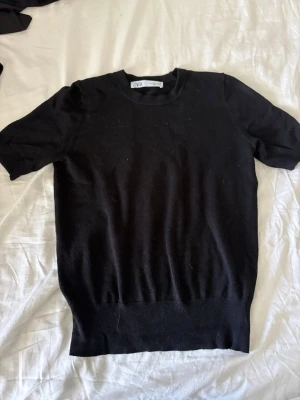 Svart kortärmad topp från Zara - En enkel och stilren svart topp från Zara med korta ärmar och rund hals. Toppen har en ribbad kant nedtill och är gjord i ett mjukt material som känns skönt mot huden. Perfekt att matcha med jeans eller kjol för en clean look.