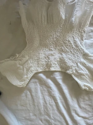 Vit smockad blus med volang - Supersöt vit blus med smockad midja och volangdetaljer upptill. Ärmlös modell med lätt struktur i tyget som ger en luftig känsla. Perfekt att styla med jeans eller kjol för en fräsch look.