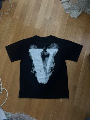Svart Vlone t-shirt med tryck - Svart t-shirt från Vlone med stort vitt V-tryck i rökdesign på ryggen och en cool fågel samt texten 'LONE' i vitt på bröstet. Klassisk passform och rund hals. Perfekt för dig som gillar streetwear och vill sticka ut.