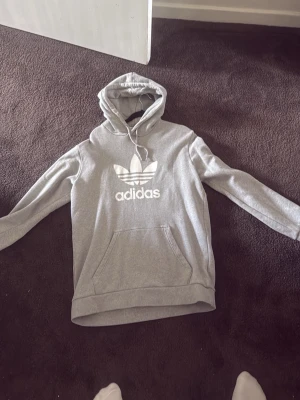 Grå Adidas hoodie med logga - Snygg grå hoodie från Adidas med stor vit Trefoil-logga och text på bröstet. Klassisk känguruficka framtill och justerbar huva med dragsko. Perfekt för en avslappnad streetstyle-look.