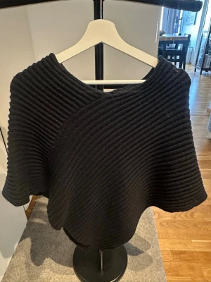 Svart stickad poncho med struktur - Snygg svart stickad poncho med markerad ribbad struktur och rundad form. Ponchon har en modern, oversized look och är perfekt att slänga över en tröja för extra värme. Materialet känns mjukt och följsamt. Enkel att styla till olika outfits. Nopprig på vissa ställen.
