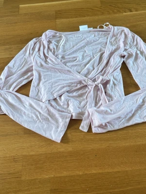 Ljusrosa omlottopp från Gina Tricot - Supersöt ljusrosa omlottopp från Gina Tricot med långa ärmar och knyt i midjan. Tunn och mjuk i materialet, perfekt för lager-på-lager eller som den är. Enkel och stilren design som passar till mycket. Har använt 1 gång 