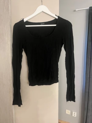 Svart långärmad topp/sweter - En enkel och stilren svart långärmad topp från gina. Modellen har en rundad, djup urringning och är figurnära i passformen. Den har en slit längst ner vid ärmarna. Perfekt att matcha med jeans eller kjol för en clean look.